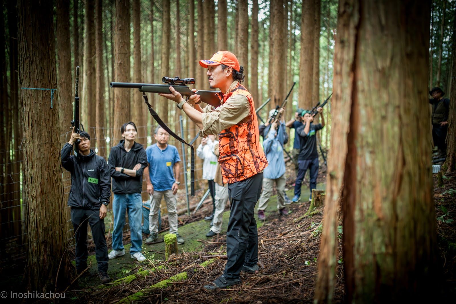 杉林の中で銃の構え方を実演する講師と、それを見つめる参加者たち