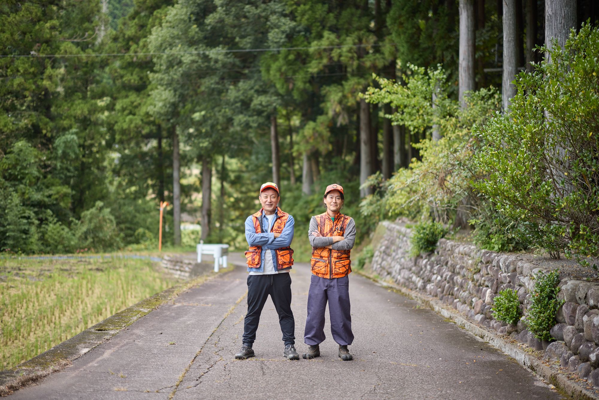 山あいの道に並んで立つ、オレンジのベストを着た講師2人